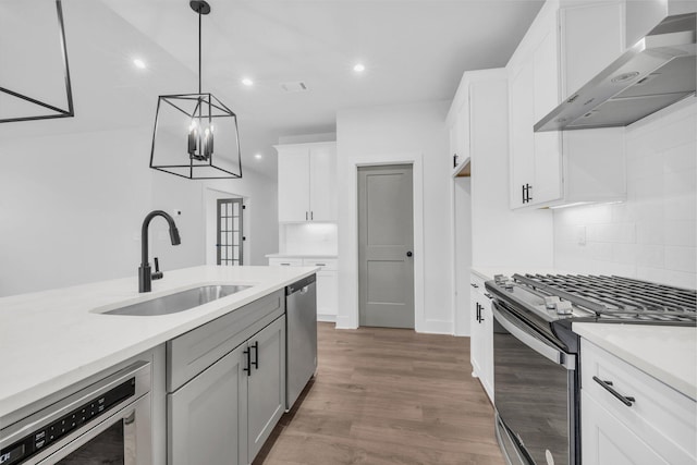 kitchen with wall chimney range hood, sink, appliances with stainless steel finishes, white cabinetry, and hanging light fixtures