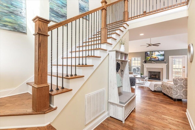 staircase featuring a fireplace, visible vents, a high ceiling, ceiling fan, and wood finished floors