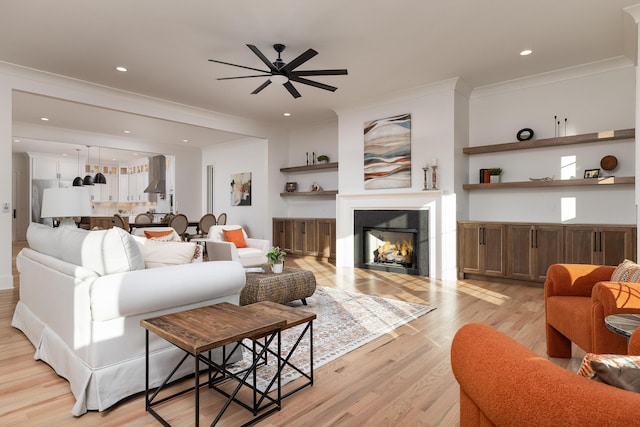 living room featuring crown molding, light wood finished floors, recessed lighting, a ceiling fan, and a warm lit fireplace