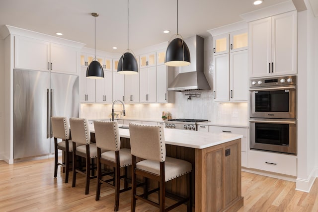 kitchen with stainless steel appliances, white cabinetry, wall chimney exhaust hood, an island with sink, and glass insert cabinets