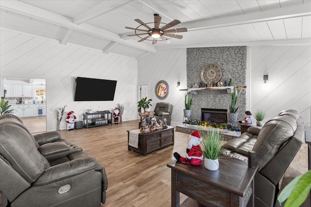 living room featuring vaulted ceiling with beams, light wood-style flooring, ceiling fan, and a stone fireplace