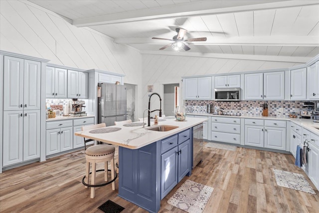 kitchen with vaulted ceiling with beams, stainless steel appliances, light wood-style floors, blue cabinetry, and tasteful backsplash