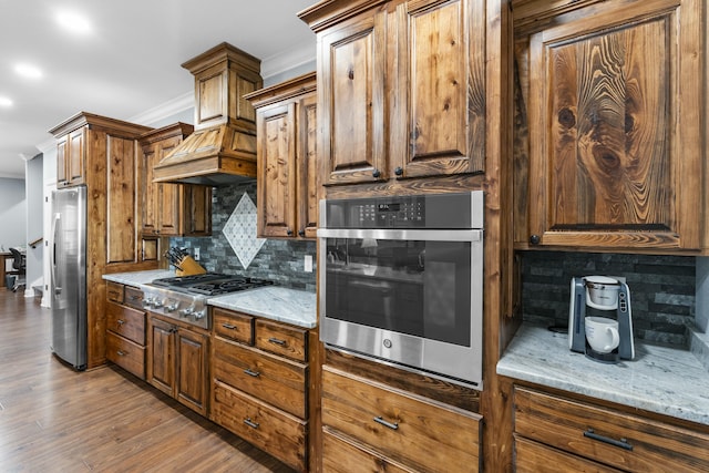 kitchen with light stone counters, backsplash, appliances with stainless steel finishes, ornamental molding, and wood finished floors