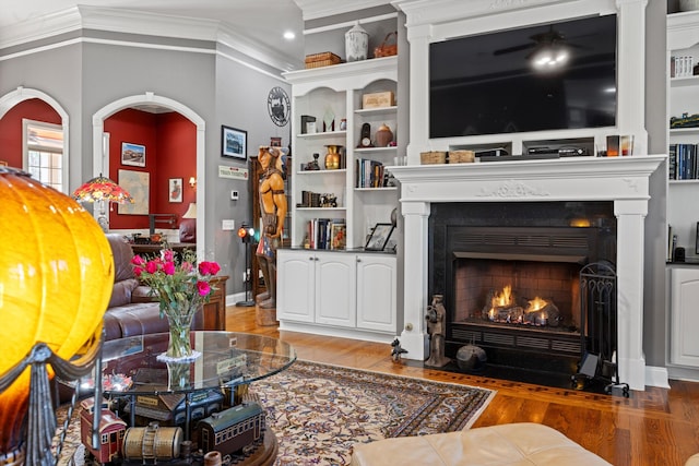 living area with built in features, a fireplace with flush hearth, ornamental molding, wood finished floors, and baseboards