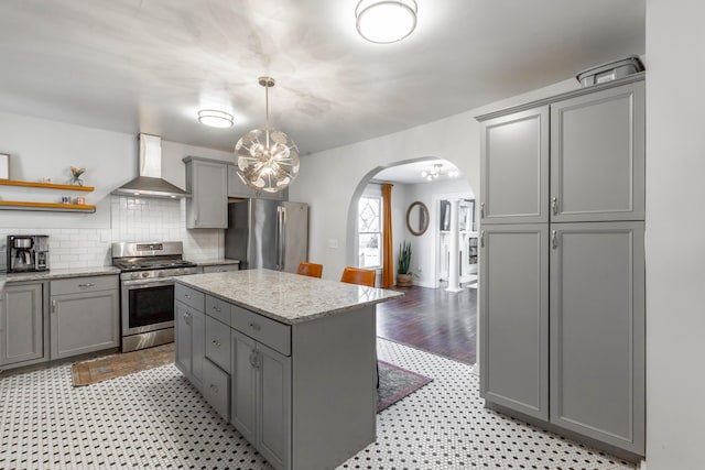 kitchen with decorative light fixtures, gray cabinets, appliances with stainless steel finishes, a kitchen island, and wall chimney range hood