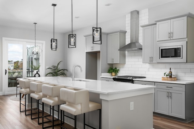 kitchen with gray cabinetry, hanging light fixtures, a center island with sink, appliances with stainless steel finishes, and wall chimney range hood