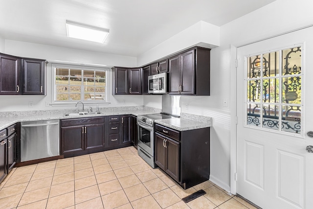 kitchen with light tile patterned floors, appliances with stainless steel finishes, dark brown cabinetry, a sink, and light stone countertops