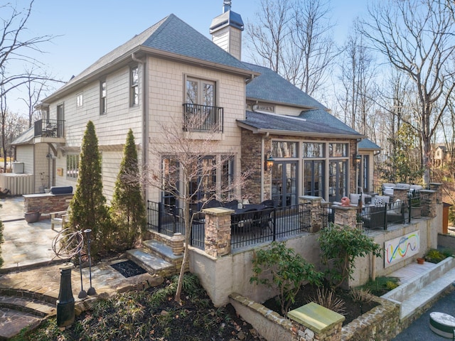 rear view of house featuring a patio, an outdoor living space, stone siding, roof with shingles, and a chimney