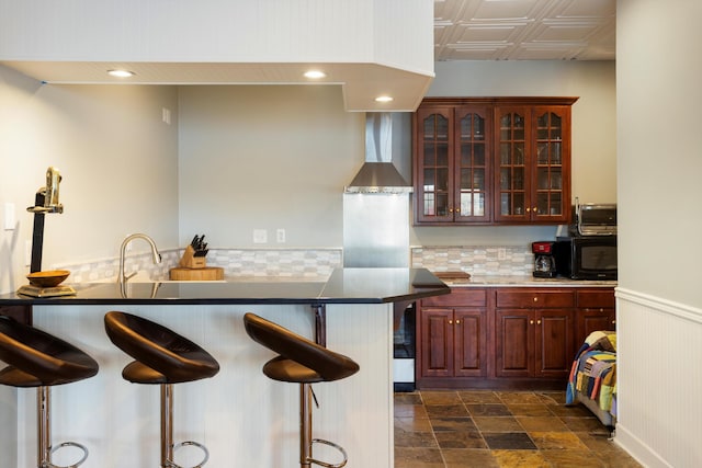 kitchen featuring a wainscoted wall, glass insert cabinets, a breakfast bar area, wall chimney range hood, and black microwave