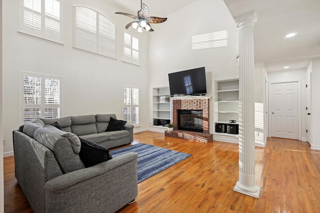 living room with a brick fireplace, a ceiling fan, wood finished floors, ornate columns, and built in shelves