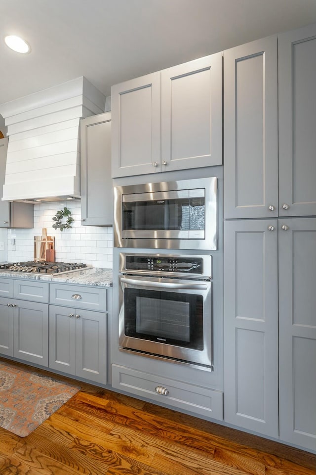 kitchen with dark wood-type flooring, appliances with stainless steel finishes, backsplash, gray cabinets, and light stone countertops