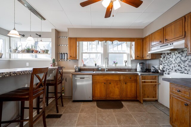 kitchen with under cabinet range hood, a sink, stainless steel dishwasher, backsplash, and brown cabinetry