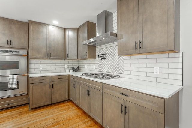 kitchen featuring decorative backsplash, wall chimney exhaust hood, stainless steel appliances, light countertops, and light wood-style floors