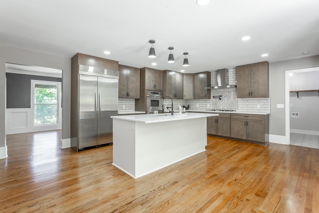 kitchen featuring light countertops, appliances with stainless steel finishes, wall chimney range hood, light wood-type flooring, and decorative backsplash