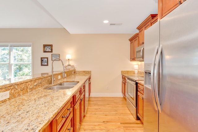 kitchen with stainless steel appliances, brown cabinetry, a sink, light stone countertops, and light wood-type flooring