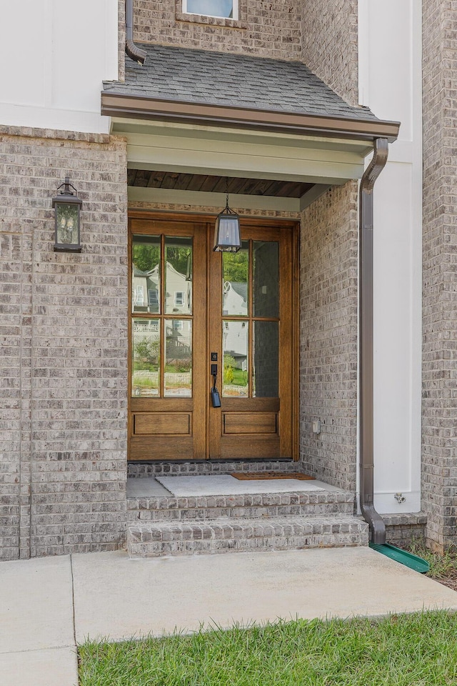 view of exterior entry with french doors, roof with shingles, and brick siding