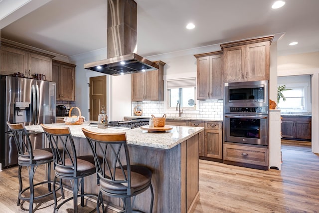 kitchen featuring island range hood, decorative backsplash, light wood-style flooring, light stone countertops, and stainless steel appliances