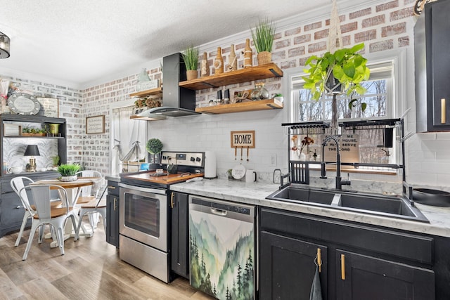 kitchen featuring stainless steel appliances, dark cabinetry, wall chimney range hood, open shelves, and a sink