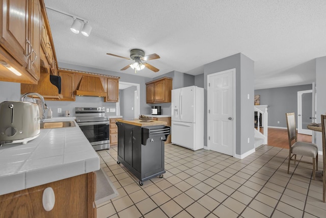 kitchen with tile countertops, white refrigerator with ice dispenser, premium range hood, a fireplace, and stainless steel range with electric stovetop