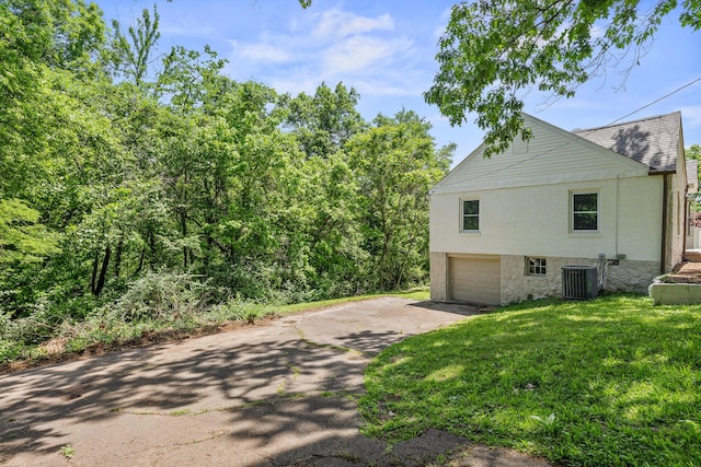 view of side of home featuring aphalt driveway, an attached garage, cooling unit, roof with shingles, and a lawn