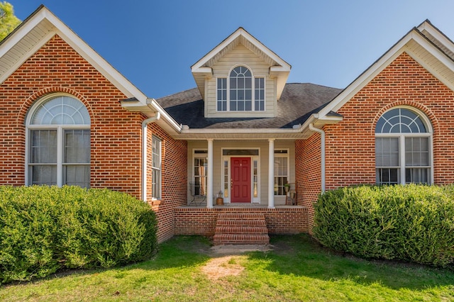 view of front facade with covered porch, a shingled roof, a front lawn, and brick siding