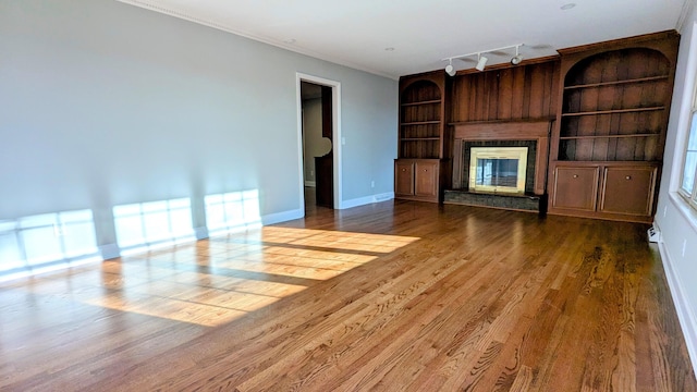 unfurnished living room featuring built in shelves, wood finished floors, baseboards, a glass covered fireplace, and rail lighting