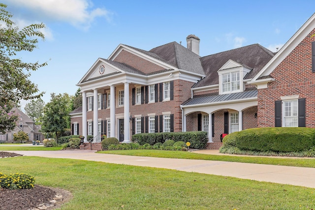 greek revival inspired property with brick siding, a chimney, a front yard, a standing seam roof, and metal roof