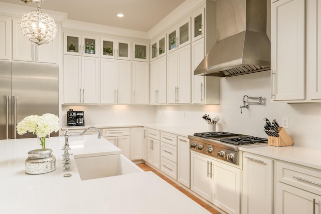 kitchen with white cabinetry, stainless steel appliances, wall chimney exhaust hood, glass insert cabinets, and hanging light fixtures