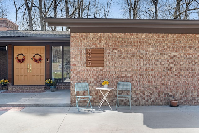 entrance to property featuring roof with shingles, a patio, and brick siding