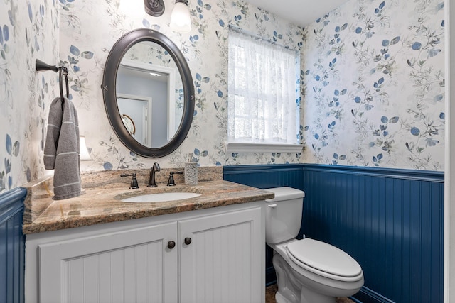 bathroom with a wainscoted wall, vanity, toilet, and wallpapered walls