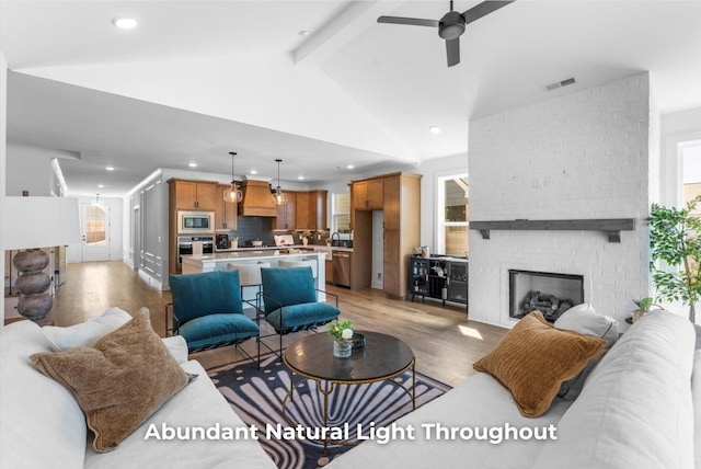 living room with a ceiling fan, visible vents, lofted ceiling with beams, a brick fireplace, and light wood-type flooring