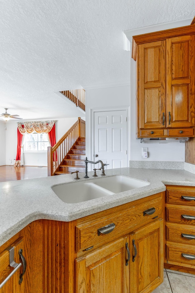 kitchen with a textured ceiling, a sink, a ceiling fan, brown cabinets, and light stone countertops