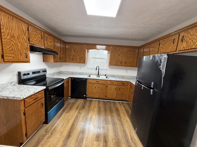 kitchen featuring brown cabinetry, a sink, light wood-type flooring, under cabinet range hood, and black appliances