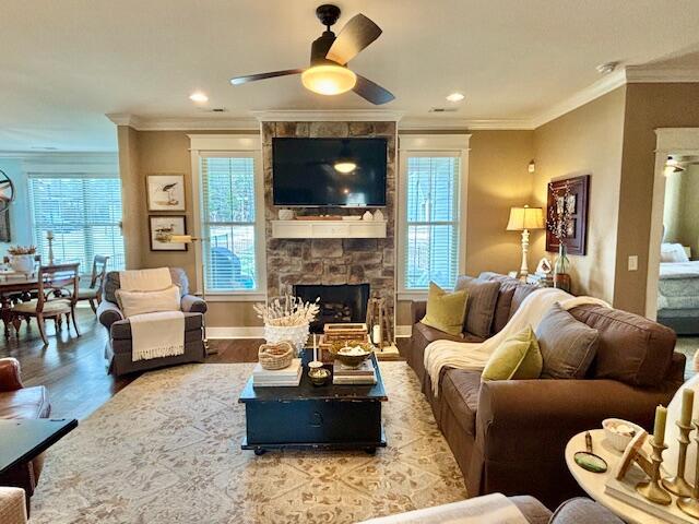 living area featuring a ceiling fan, wood finished floors, baseboards, ornamental molding, and a stone fireplace