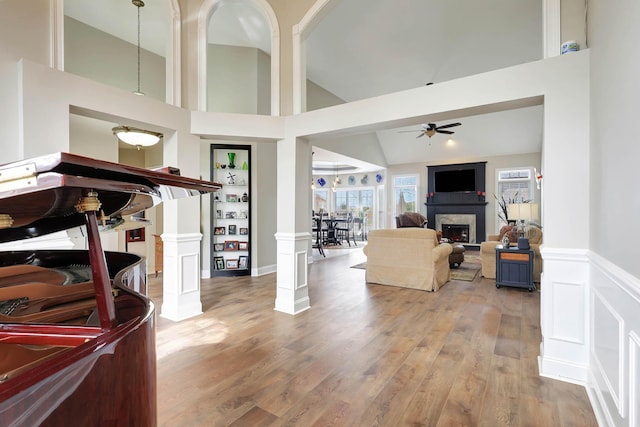 living area featuring a ceiling fan, wood finished floors, a wainscoted wall, ornate columns, and a fireplace