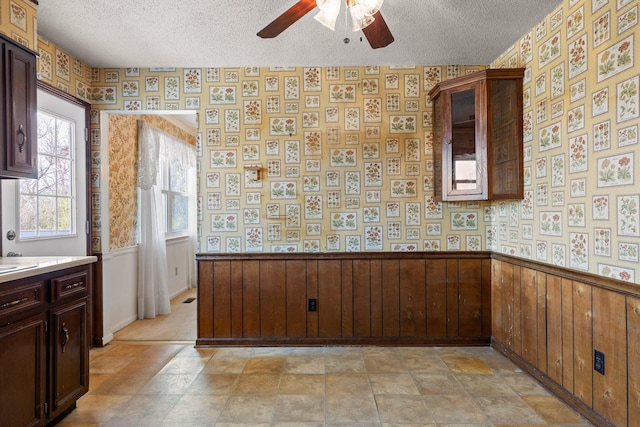 kitchen featuring a wainscoted wall, a textured ceiling, ceiling fan, and wallpapered walls