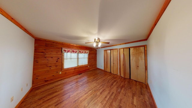 unfurnished bedroom with ceiling fan, light wood-style floors, wood walls, crown molding, and two closets