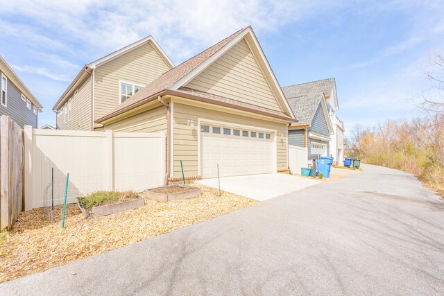 view of side of property featuring an attached garage, concrete driveway, roof with shingles, and fence