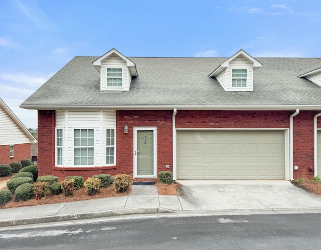 new england style home with brick siding, driveway, an attached garage, and roof with shingles