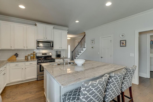 kitchen with a kitchen island with sink, a sink, ornamental molding, stainless steel appliances, and tasteful backsplash