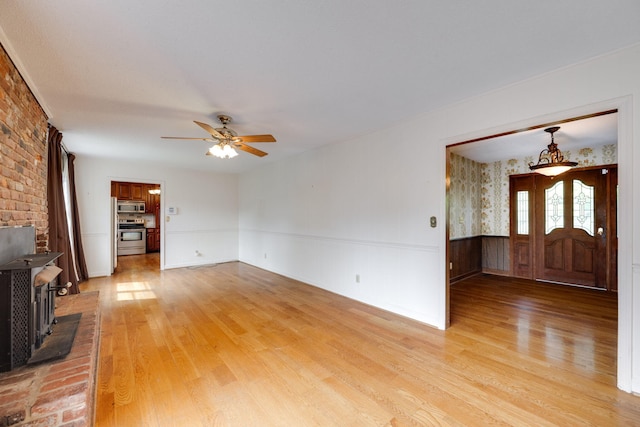 unfurnished living room featuring a wood stove, light wood-style floors, a wainscoted wall, and ceiling fan