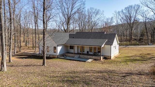 back of house featuring a forest view, a lawn, a shingled roof, and a patio