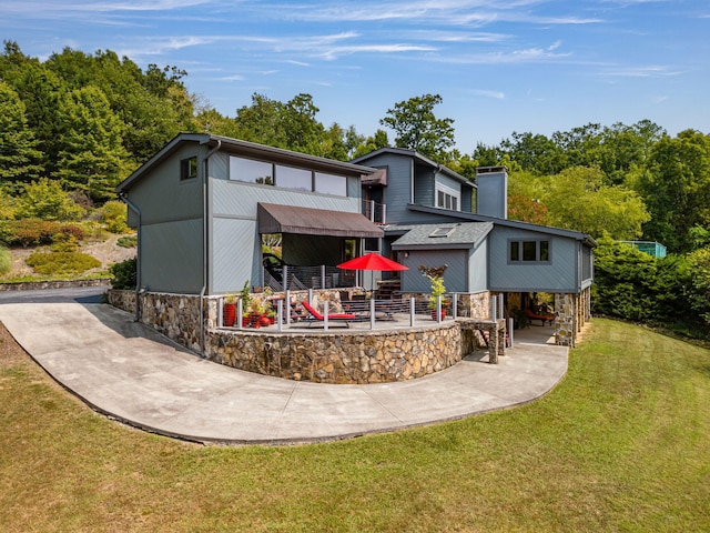 back of house with concrete driveway, a lawn, a chimney, stone siding, and a patio