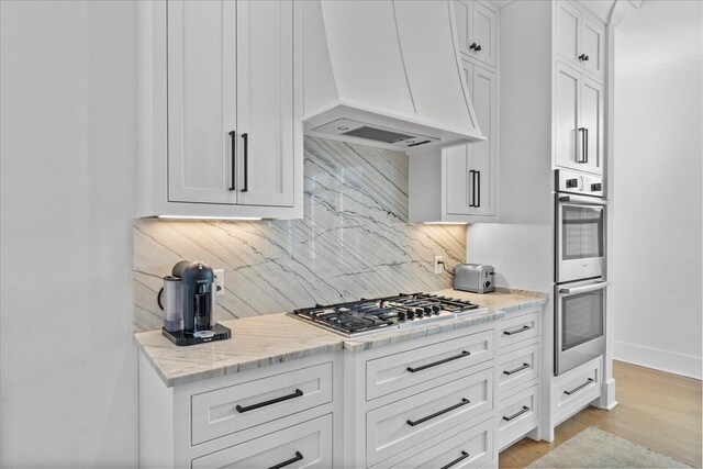 kitchen featuring premium range hood, backsplash, white cabinetry, light stone countertops, and stainless steel appliances