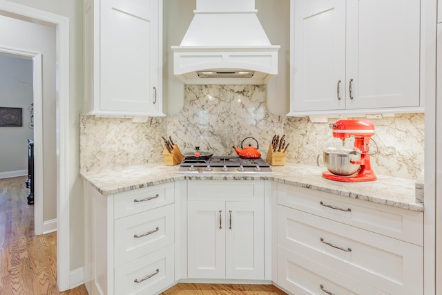 kitchen with decorative backsplash, stainless steel gas cooktop, premium range hood, white cabinets, and light stone counters