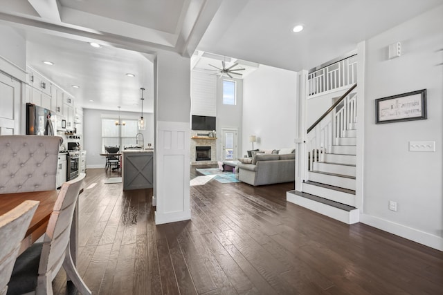 living area with ceiling fan, a stone fireplace, stairway, dark wood-style flooring, and baseboards