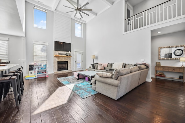 living area featuring ceiling fan, a towering ceiling, a stone fireplace, hardwood / wood-style floors, and baseboards
