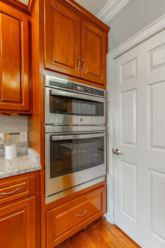 kitchen with light stone counters, decorative backsplash, brown cabinets, double oven, and ornamental molding