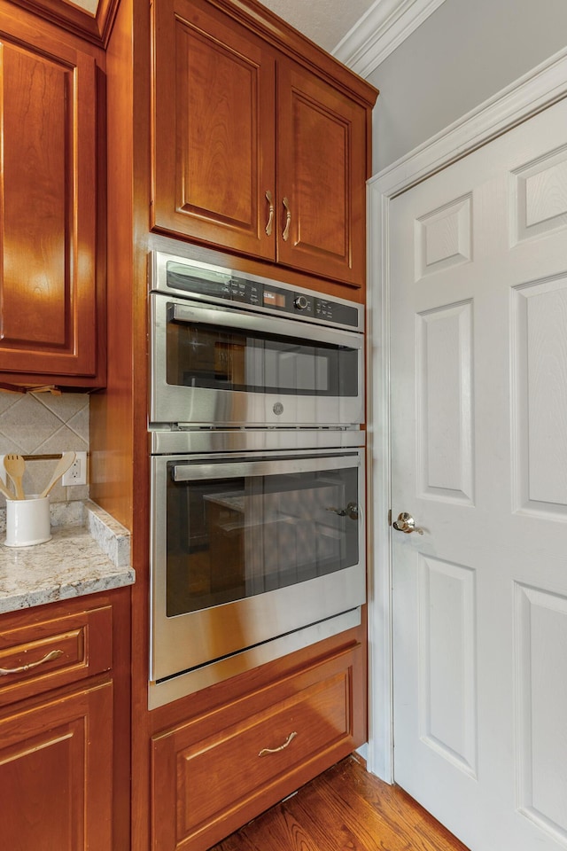 kitchen featuring tasteful backsplash, light stone counters, brown cabinets, double oven, and ornamental molding