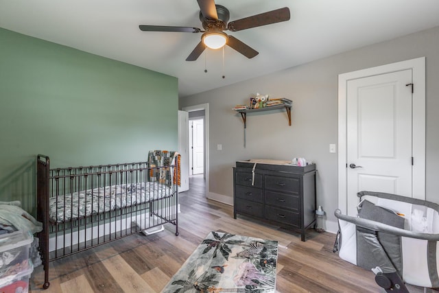 bedroom with baseboards, a ceiling fan, a nursery area, and wood finished floors